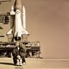 A white woman in her 30s sits on the launchpad, with the space shuttle (configured for launch) behind her. She has long brown hair and is looking to the side.