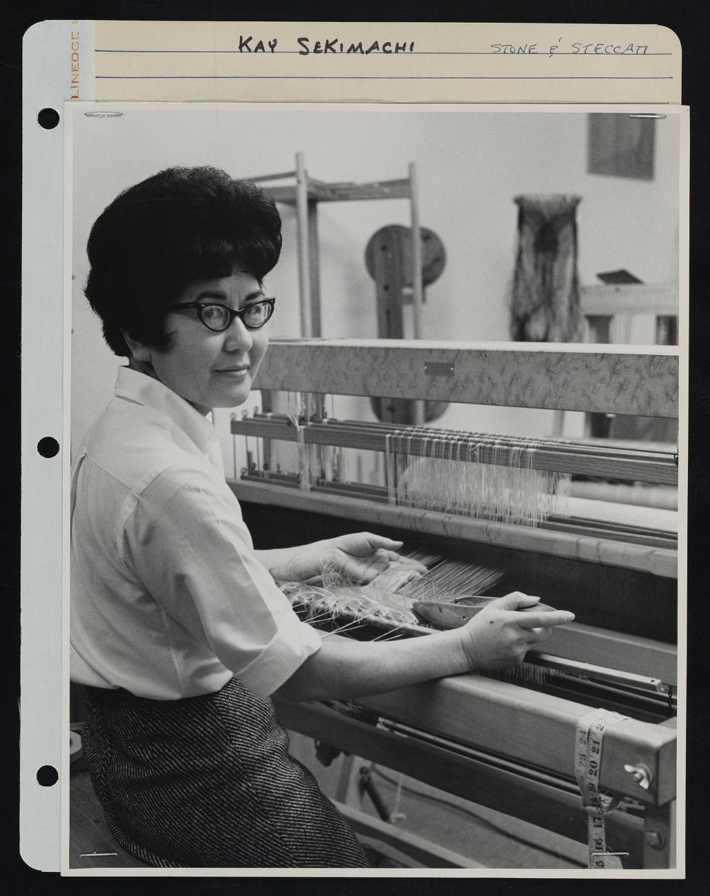 Black and white image of a woman sitting at a loom, looking at the camera