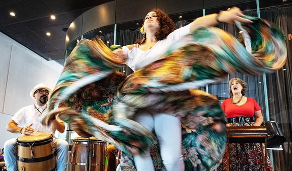 A person dances, turquoise and brown skirt flying and twisting around them, with a drummer and singer performing on the sides.