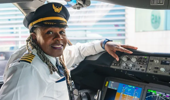 A 65-year-old Black woman sits in the left seat of an airliner cockpit. She is smiling and wearing an airline captain's uniform, including a white shirt and a hat.