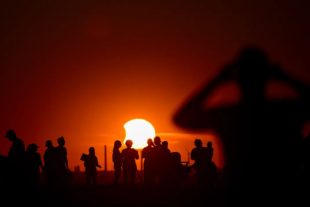 a low-hanging sun with a small bit of it covered by the moon at left, behind a crowd of people in silhouette