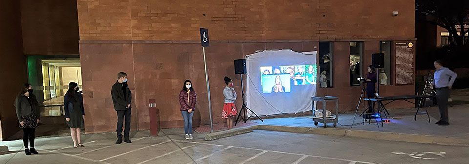 Students stand in front of a brick wall with a sheet hanging on part of it to create a drive in movie setting.