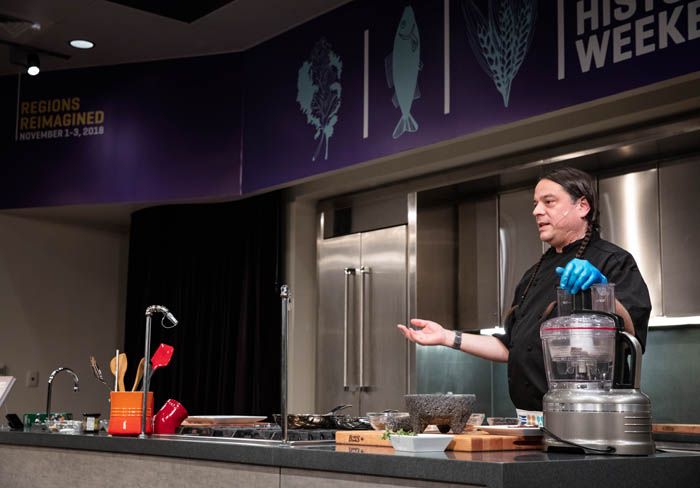 Chef Sean Sherman cooking in our demonstration kitchen, one hand on a food processor and one hand explaining