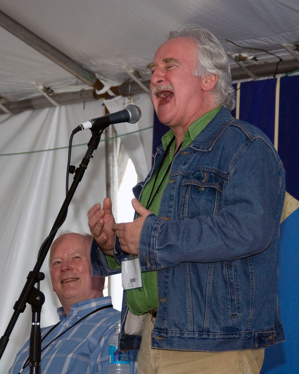 A man in a kelly green dress shirt and blue denim jacket sings or shouts, mouth wide and eyes shut, into a microphone on stage under a festival tent. Another man seated beside him looks up at him and smiles.