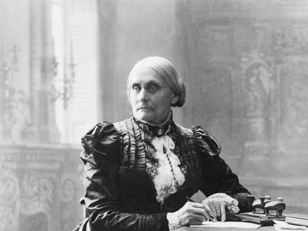 A black and white photograph of a woman with a tight bun, wearing a ruffled black dress with a white neckpiece, sitting a desk and in the middle of writing a letter, not smiling 