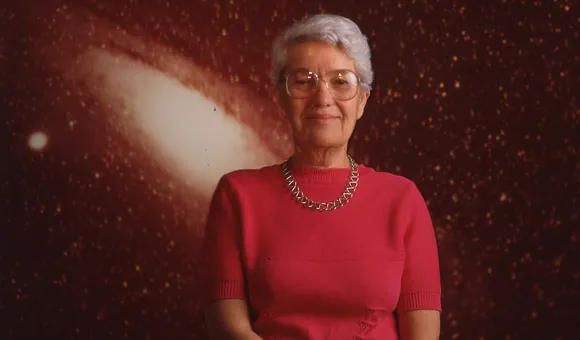 Vera Rubin stands against a backdrop of a star-filled galaxy with her hands loosely clasped in front of her.