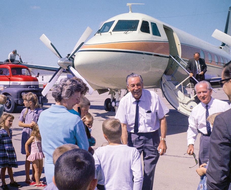 Walt Disney, an senior man in a white button up short-sleeved shirt, with a tie and slacks, greets a small crowd of people in front of his airplane, which is large enough it extends out of frame and is white with orange and blue stripes.