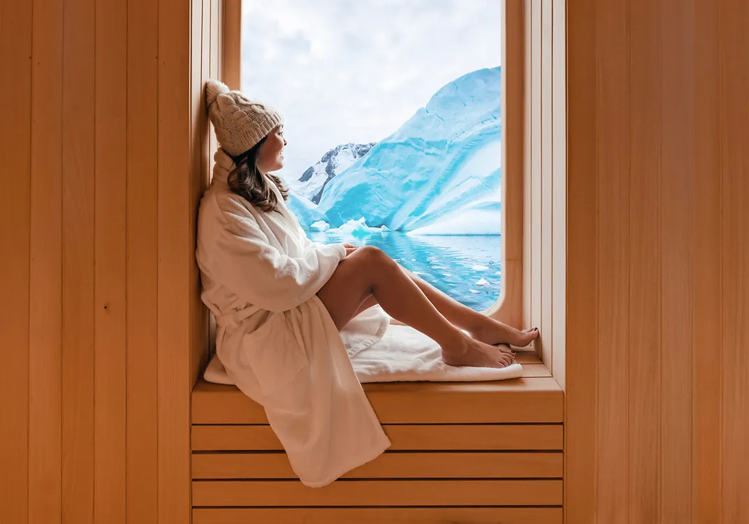 A person in a white bathrobe and knit hat sits on a wooden bench aboard the World Navigator, looking out a large window at blue glaciers and calm Antarctic waters.