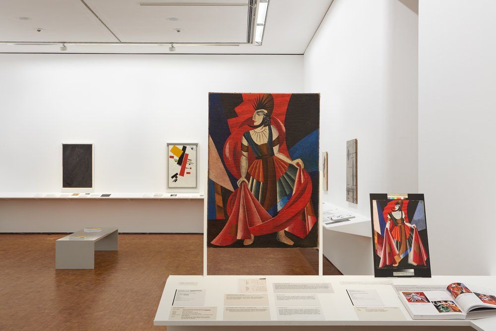 Stark white museum walls with canvases hanging on the walls at intervals; in foreground, a large red and black abstracted composition of a woman in a dress; to the right, a smaller version of the same work
