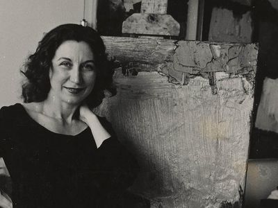 Grayscale photograph of smiling woman dressed in a black top and pants with her right hand resting on her hip and her right hand next to her neck standing in front of an abstract painting on an easel.