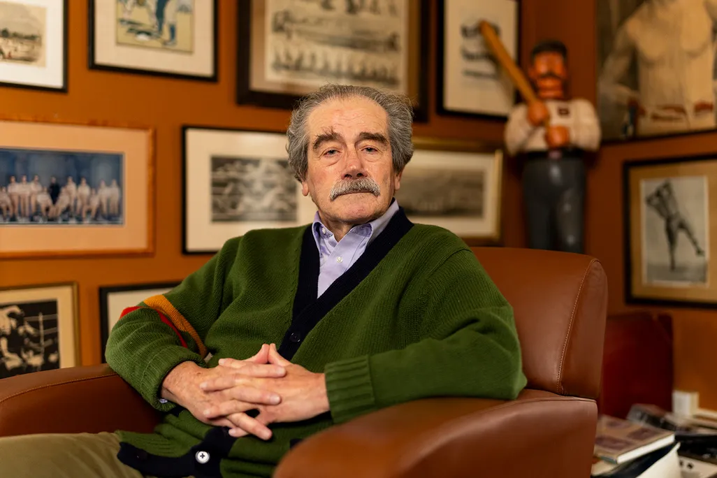 A man in a green sweater sits in front of a wall of hanging photographs