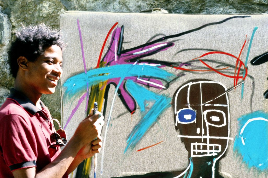 Jean-Michel Basquiat, smiling, stands in front of one of his works in progress. It's sunny outside, and he wears a red button down shirt