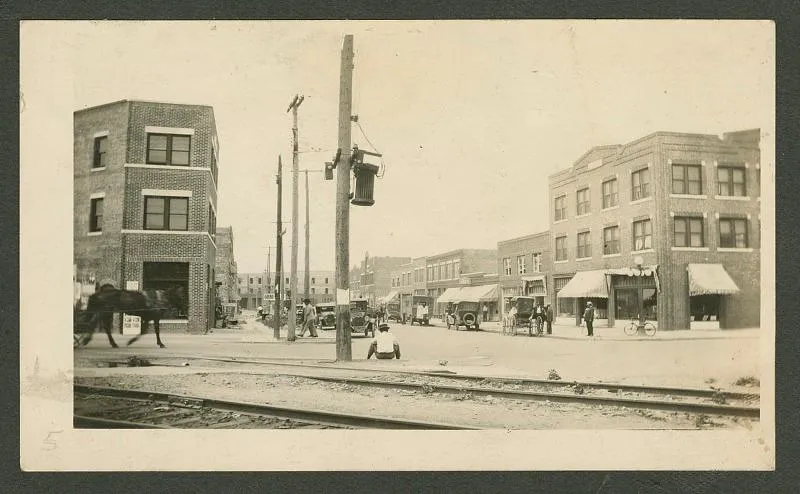 A yellow-toned black and white image of a cross-street, with a horse and carriage on the left, a Black person sitting in center frame, and many businesses and storefronts lining a city street