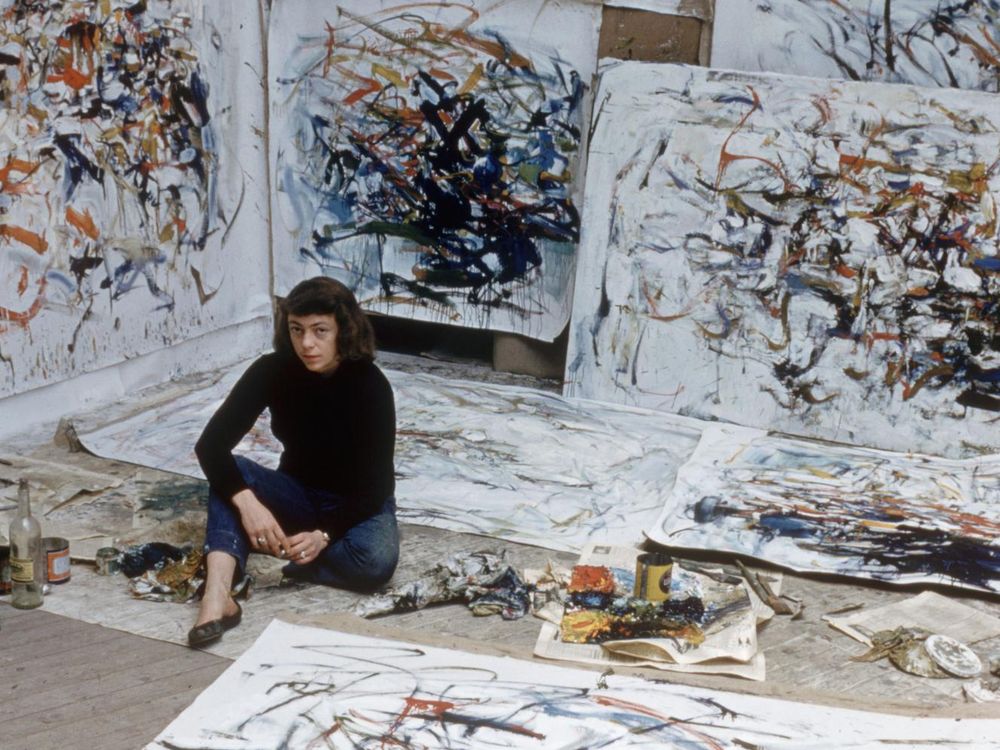 A view of Mitchell, wearing a black turtleneck, seated amid an explosion of white canvases with markings