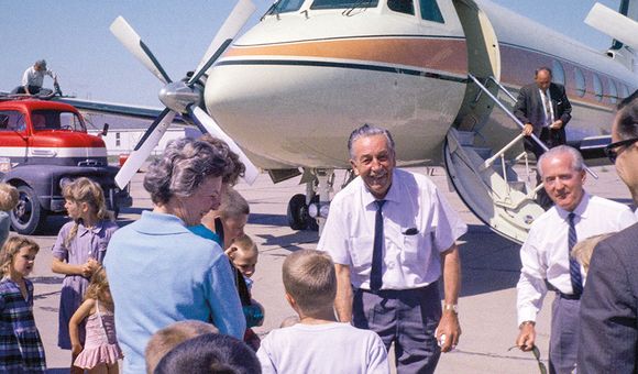 Walt Disney, an senior man in a white button up short-sleeved shirt, with a tie and slacks, greets a small crowd of people in front of his airplane, which is large enough it extends out of frame and is white with orange and blue stripes.