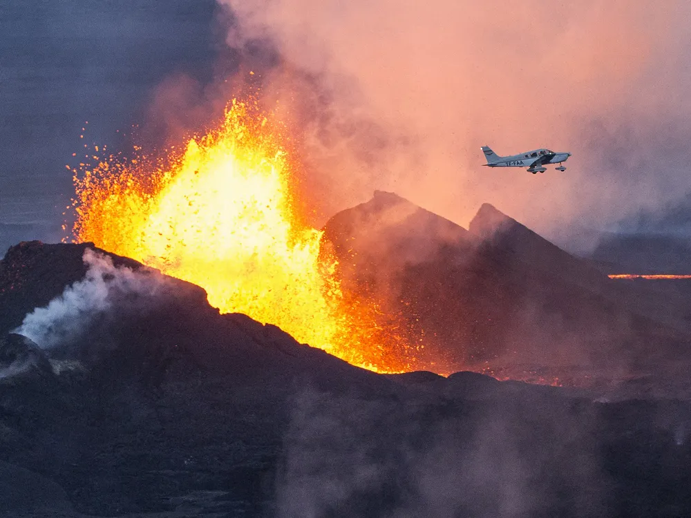 Red hot molten lava and thick dark dust spew from the ground while a small white. single-propeller aircraft flies overhead.