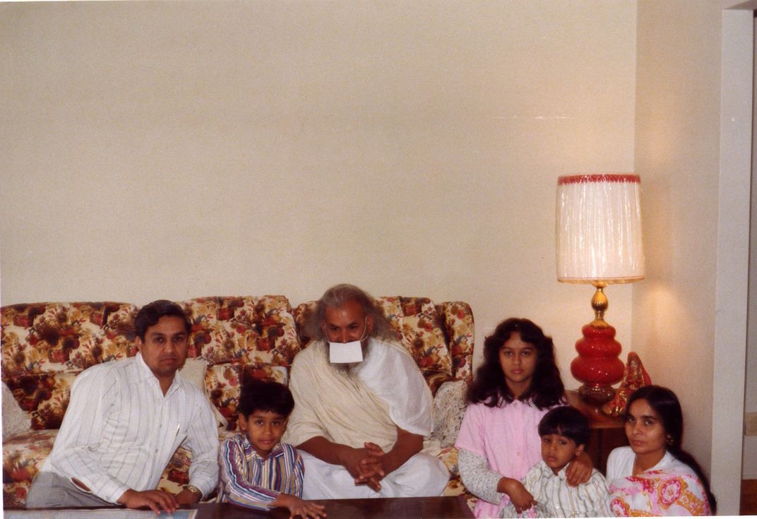 Old family photo with the Jain family and an older man posing, sitting on and around a beige floral-patterned couch. The older man is wearing a white face mask.