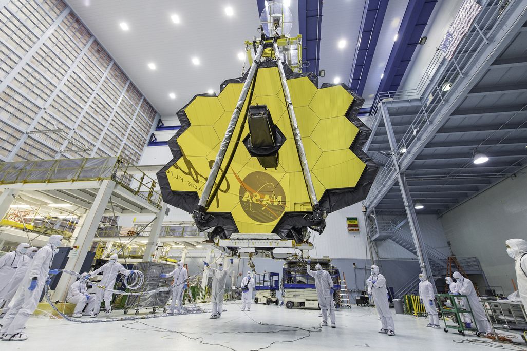 An image of the James Web Telescope in a NASA laboratory. Technicans are seen standing under it while the honey come shaped telecope mirror hangs above. The NASA logo is seen reflected in the mirrors.