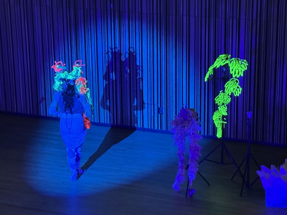 Armando Lopez-Bircann wears a cobalt blue bodysuit and skirt on a stage bathed in blue light. They have a neon green, blue, and pink headdress on and face away from the audience, with in front of their shadow. To the right are props including a neon green