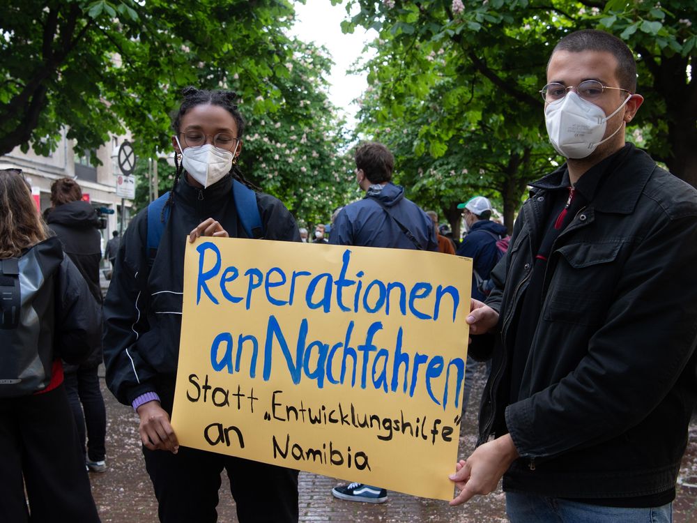 Two people in masks hold a yellow sign with blue and black handpainted letters, standing in a city street in front of trees and other protesters