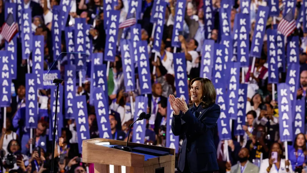 Kamala Harris looks out onto the crowd behind a podium. Audience members hold a variety of signs that say "Kamala"