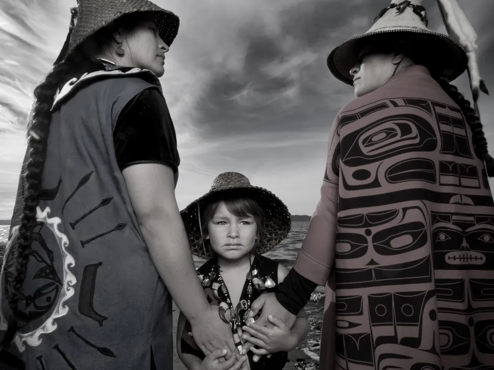 Darkfeather Ancheta, Eckos Chartraw-Ancheta, and Bibiana Ancheta, Tulalip, Washington. Darkfeather, is pictured with her sister Bibiana and nephew Eckos at the edge of Tulalip Bay. They are wearing traditional regalia prepared for their annual Canoe Journey. Every year, upwards of 100 U.S. tribes, Canadian First Nations and New Zealand canoe families will make “The Journey” by pulling their canoes to a host destination Tribe. Canoe families pull for weeks, and upon landing participate in several days and nights of “protocol”, a celebration sharing traditional knowledge, ancestral songs, and sacred dances. Photo by Matika Wilbur for Project 562. Courtesy of the Artist. 