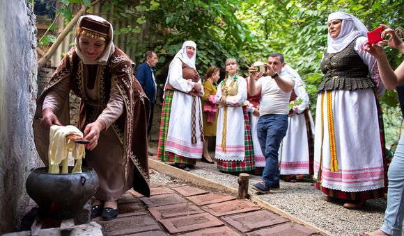 A woman wearing traditional Armenian dress bends down to tend to an outdoor stove while preparing a meal.