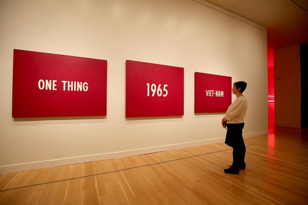 A photograph of a woman standing in front of a three panel painting that has words that say, "On thing, 1965, Viet-nam." 