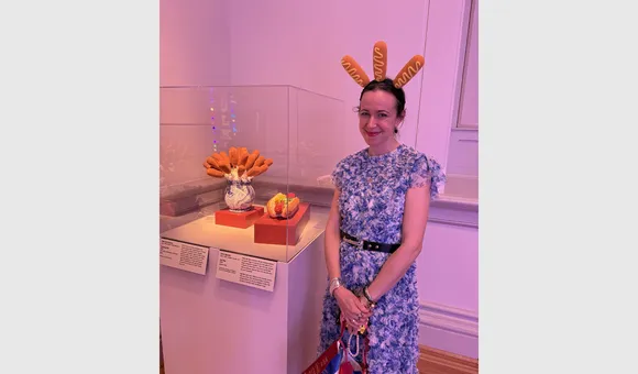 Sarah Rogers Morris wearing a headband with three corndogs poking out stands behind an exhibition case with a vase containing corndogs and a ceramic hot dog.