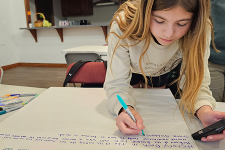 A student leans over a table, writing on a large poster board with a marker during a classroom activity. Surrounding the workspace are colorful pens, papers, and notes, while additional tables, chairs, and a kitchen area with cabinets and a counter appear