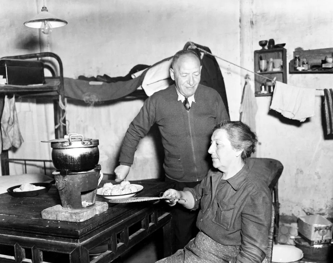 A black and white image of a woman sitting and heating a steaming pot on a small heated pot, with a man behind her and a bunk bed visible