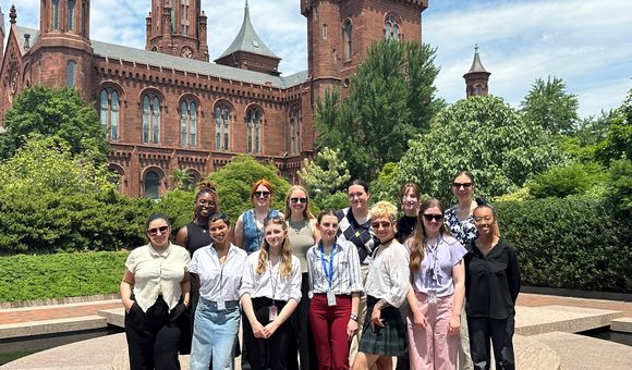 A group of thirteen people stand together in front of green shrubs and the Smithsonian Castle.