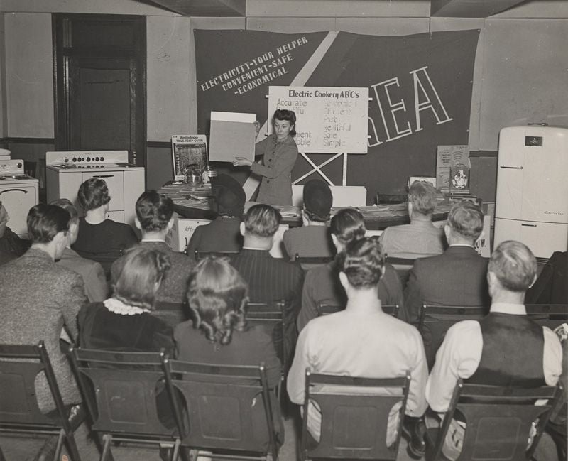 A woman delivers a presentation in front of a large group of seated people. The stage is filled with various electrical appliances, as well as a large banner with the text, "Electricity, your helper, convenient, safe, economical"