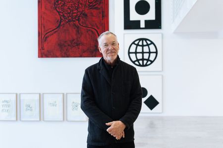 Artist Matt Mullican wearing glasses and a black jacket with his hands folded in front of his body, standing in front of six artworks, with his body partially hiding them.