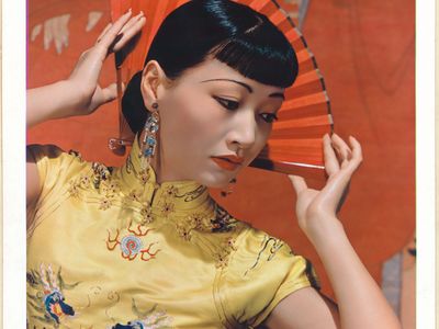 Portrait of Anna May Wong. Wong wears a yellow dress and gazes to the side of the frame. She delicately holds a red folding fan behind her head.
