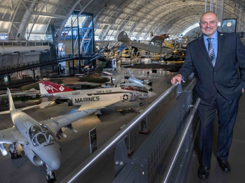 A white man in his mid-50s stands on a mezzanine overlooking several military jet airplanes parked on the concrete floor of a large hangar. The man is smiling and wearing a dark gray suit, blue shirt, and tie.