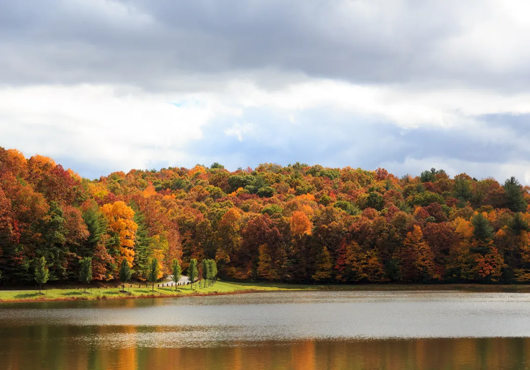 A calm lake reflects vibrant fall foliage at The Resort at Glade Springs. Hillsides covered in shades of orange, red and gold rise beyond the water beneath a sky filled with layered clouds.