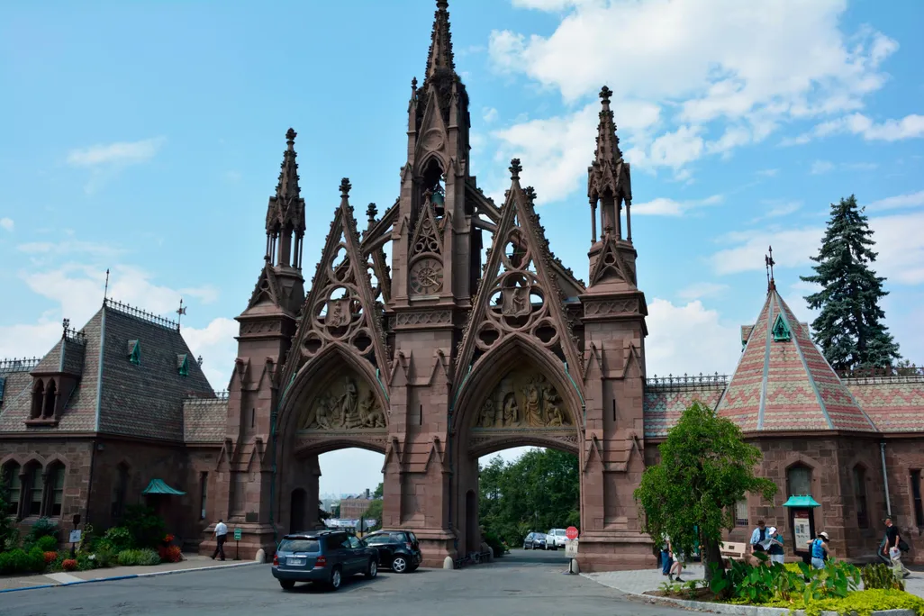 The Gothic revival entrance of Green-Wood Cemetery, two tall pointed archways and a third tall spire in the middle, in a reddish brick color with a blue sky behind