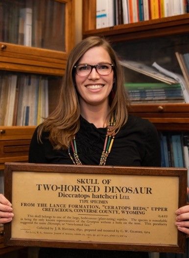 Diana Marsh holds a wood exhibit label from the Smithsonian's early fossil hall with bookshelves behind her. 