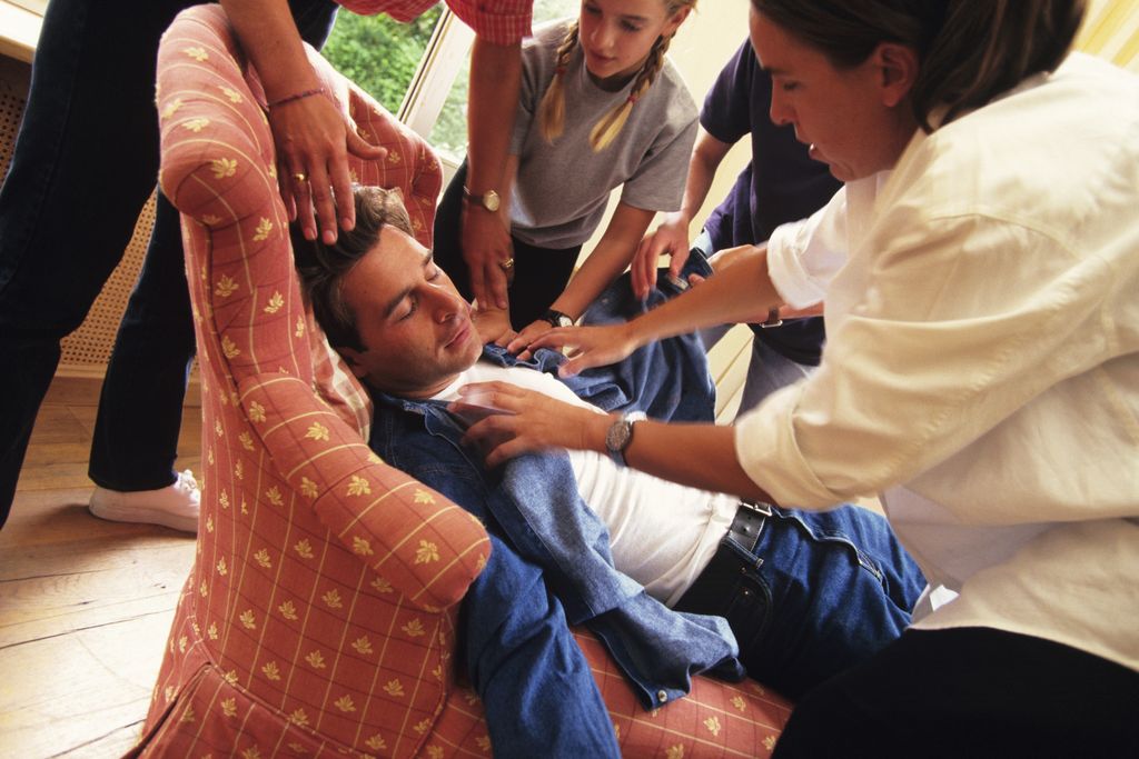 a man fainted in an armchair with four people reaching out to help him