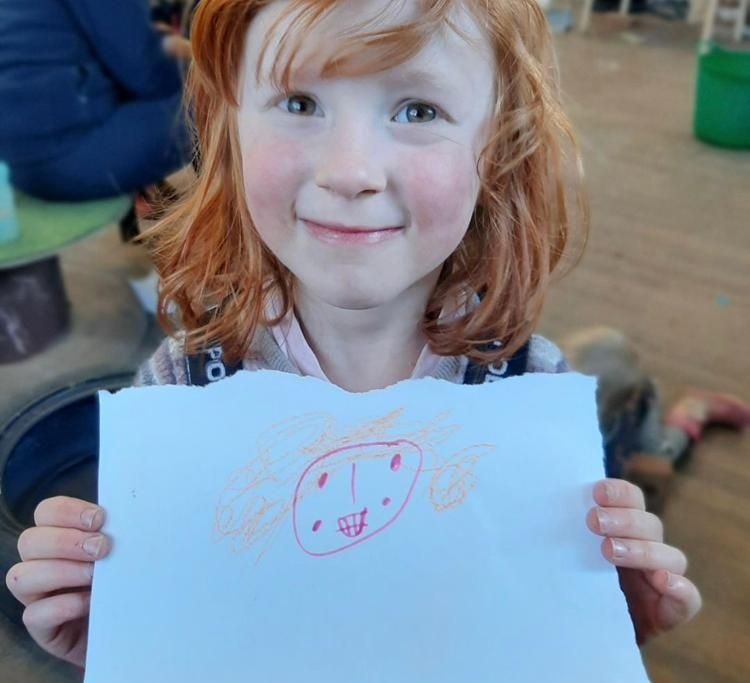 A pale, red-headed girl with bangs smiles and holds a piece of paper in front of herself, with what looks like a drawing of a person's smiling face and orange curly hair