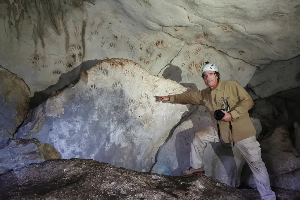 A man wearing boots and a white helmet stands in front of a large rock and points to handprints, which are reddish or white against black shadows and cover the sloping wall of the cave