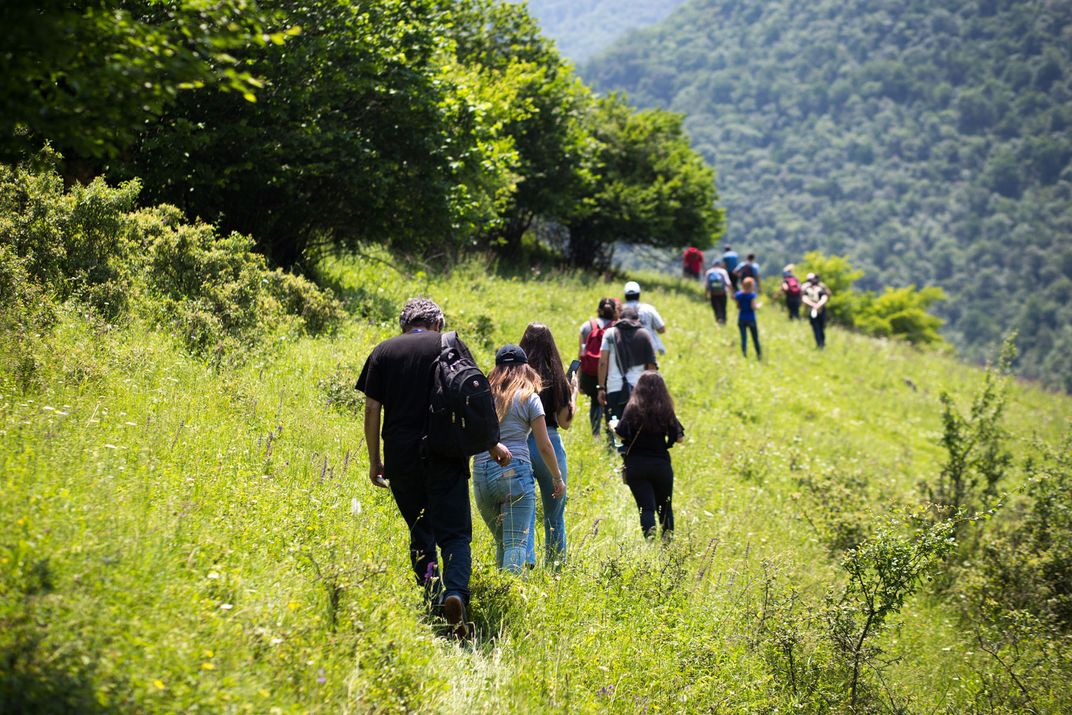 A group of hikers walk through a field towards a mountain, their backs facing away from the camera.