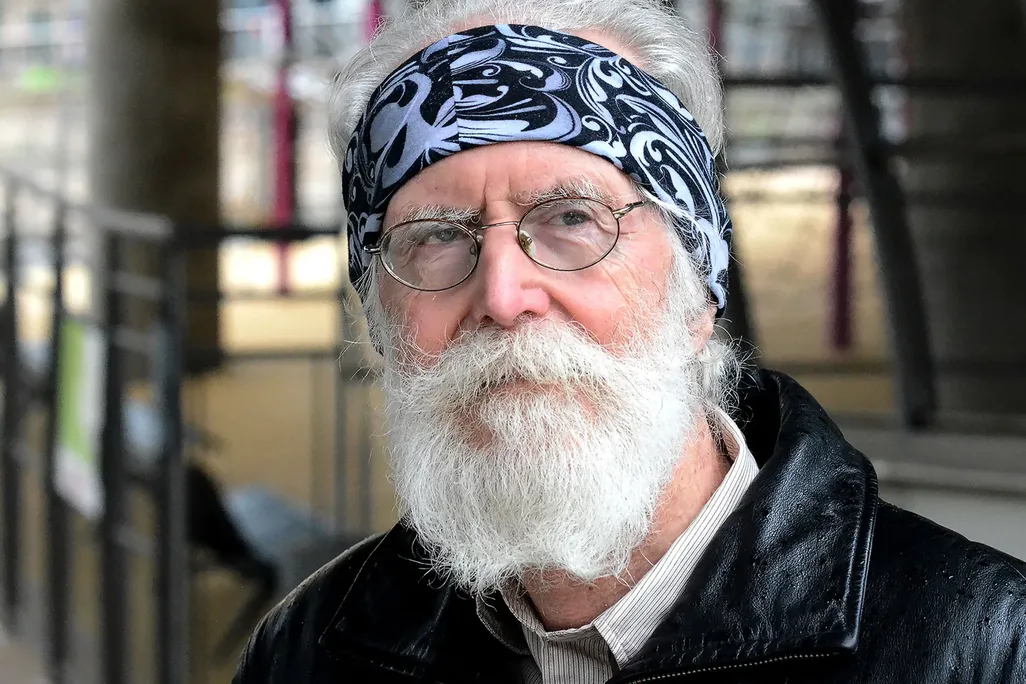 A close-up Michel Talagrand, who wears a black leather jacket, glasses, and a blue bandana wrapped around his forehead, looking to the right of the camera.