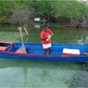 A woman in a red shirt stands on a wooden blue canoe beside a mangrove shoreline, unwinding a fishing net.