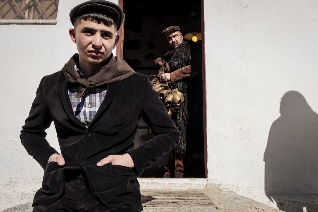 Mamuthone Matteo Gungui is dressed in the first layer of his costume: a velvet jacket, a checked shirt and a head kerchief. Behind him, an older Mamuthone is already wearing his sheepskin cloak and is about to strap on his heavy bells.