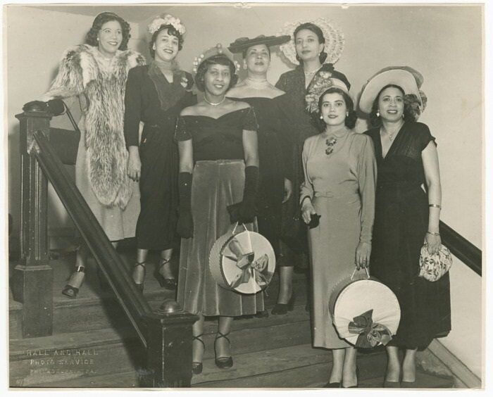 A black-and-white photograph of seven women standing on a flight of stairs. Mae Reeves stands to the farthest right on the lowest step and wears a white hat and floral patterned handbag.