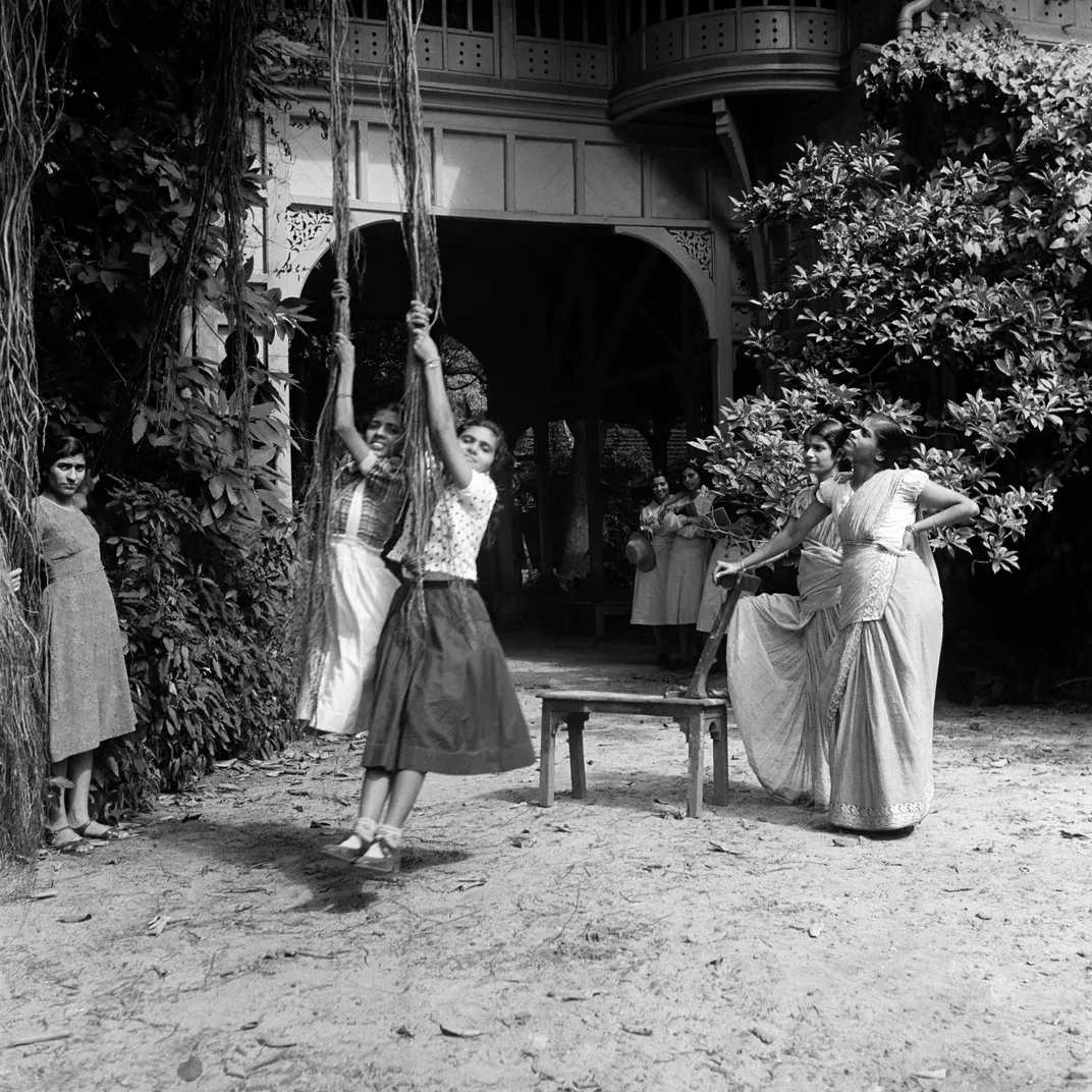 A black and white image of school girls in saris and uniforms playing on rope swings in a courtyard