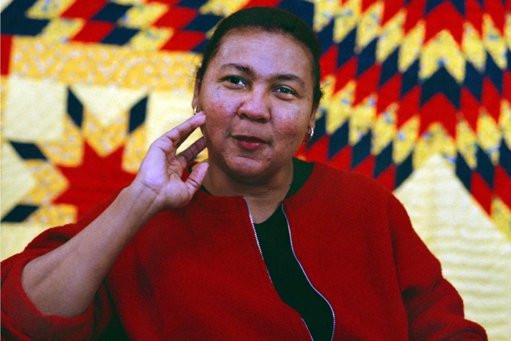hooks, a Black woman in a red blazer, sits in front of a red, yellow and black quilt with her hand resting lightly on her chin and a small smile