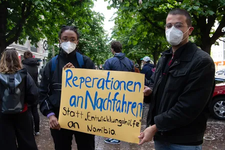 Two protesters hold a sign reading "Reparations to descendants instead of 'development aid' to Namibia" at a demonstration in Berlin on May 28. That day, the German foreign minister formally acknowledged the Herero and  Nama genocide and promised €1.1 billion in infrastructure aid—but stopped short of labeling the effort "reparations."